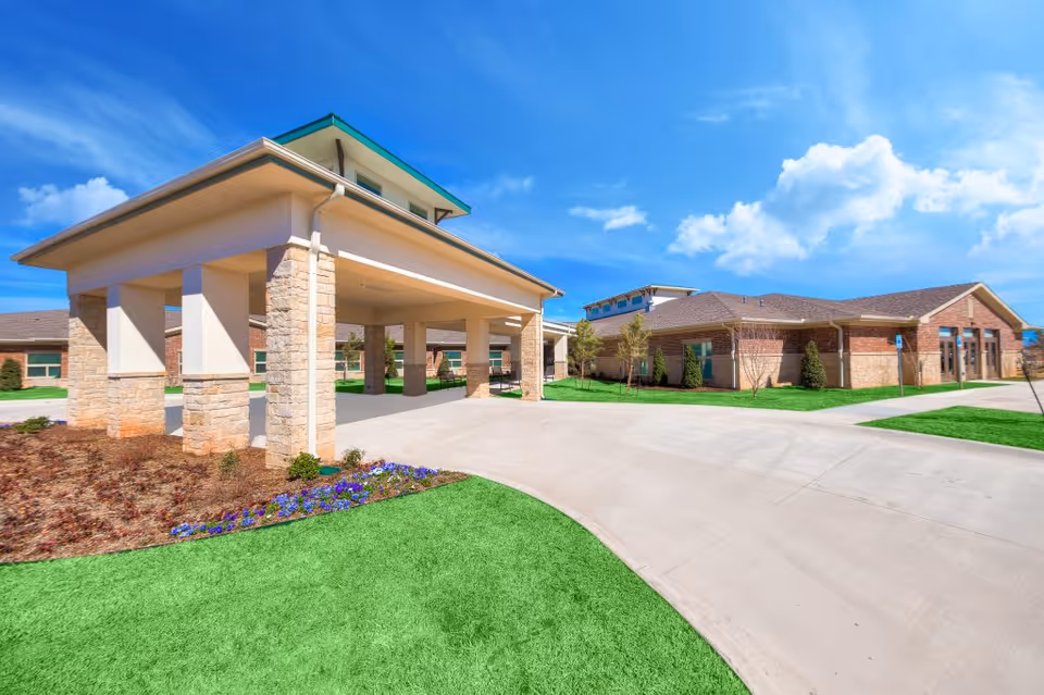 Exterior view of Accel at Crystal Park facility showing a covered entrance with stone pillars, a driveway, green grass, and a clear blue sky with some clouds.