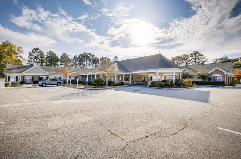 Exterior view of a single-story senior living facility building with a large covered entrance, surrounded by trees and a mostly empty parking lot under a partly cloudy sky.