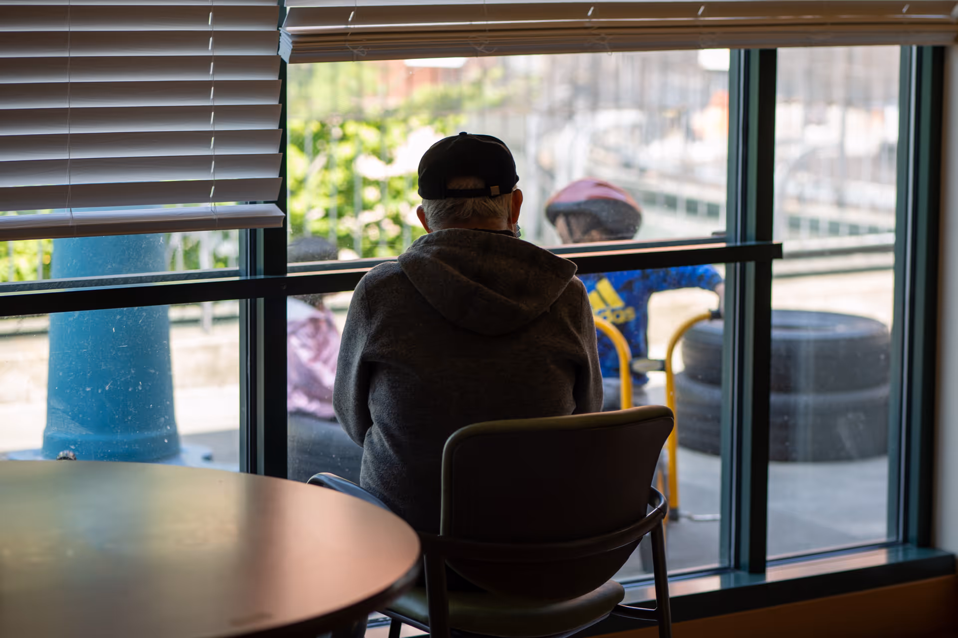An elderly person wearing a black cap and gray hoodie sitting on a chair indoors facing a large window. Outside the window, a child wearing a pink helmet and blue jacket is visible, along with some tires and outdoor play equipment.