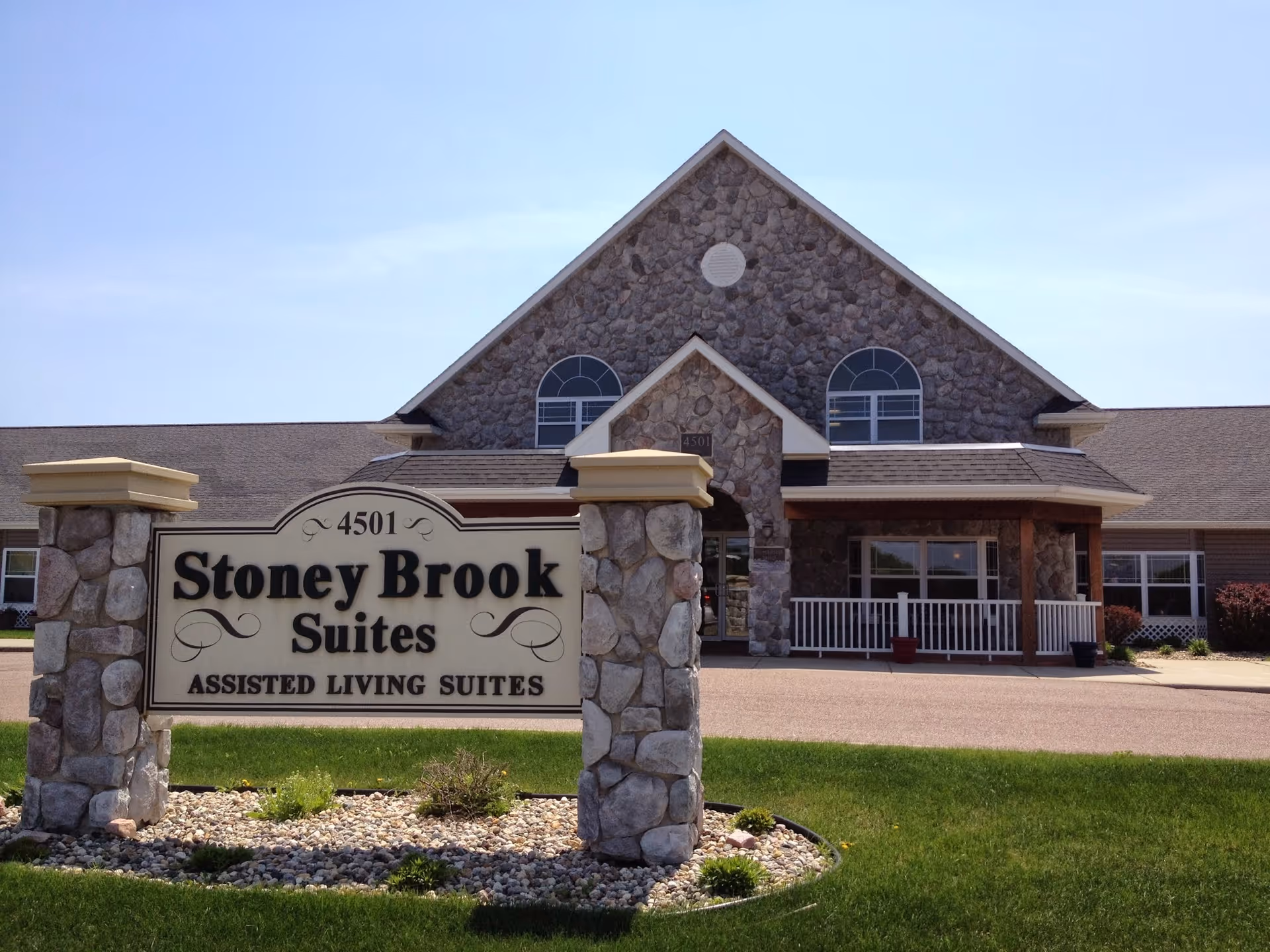Front exterior view of Stoney Brook Suites Assisted Living building with a stone facade and a sign in front displaying the facility name and address 4501. The building has a peaked roof, arched windows, and a small porch with white railings. The surrounding area includes a well-maintained lawn and landscaping with rocks and small plants.