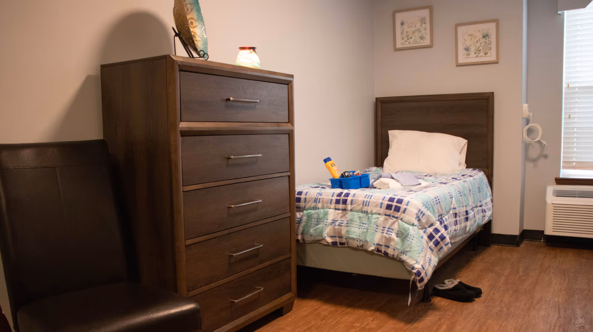 A small bedroom with a single bed covered in a blue and green plaid blanket, a white pillow, and a blue tray with personal items on the bed. Next to the bed is a tall wooden dresser with five drawers and a decorative item on top. A black leather chair is positioned beside the dresser. The room has light-colored walls, two framed pictures hanging above the bed, a window with blinds, and a heating/cooling unit below the window. A pair of black slippers is on the wooden floor near the bed.