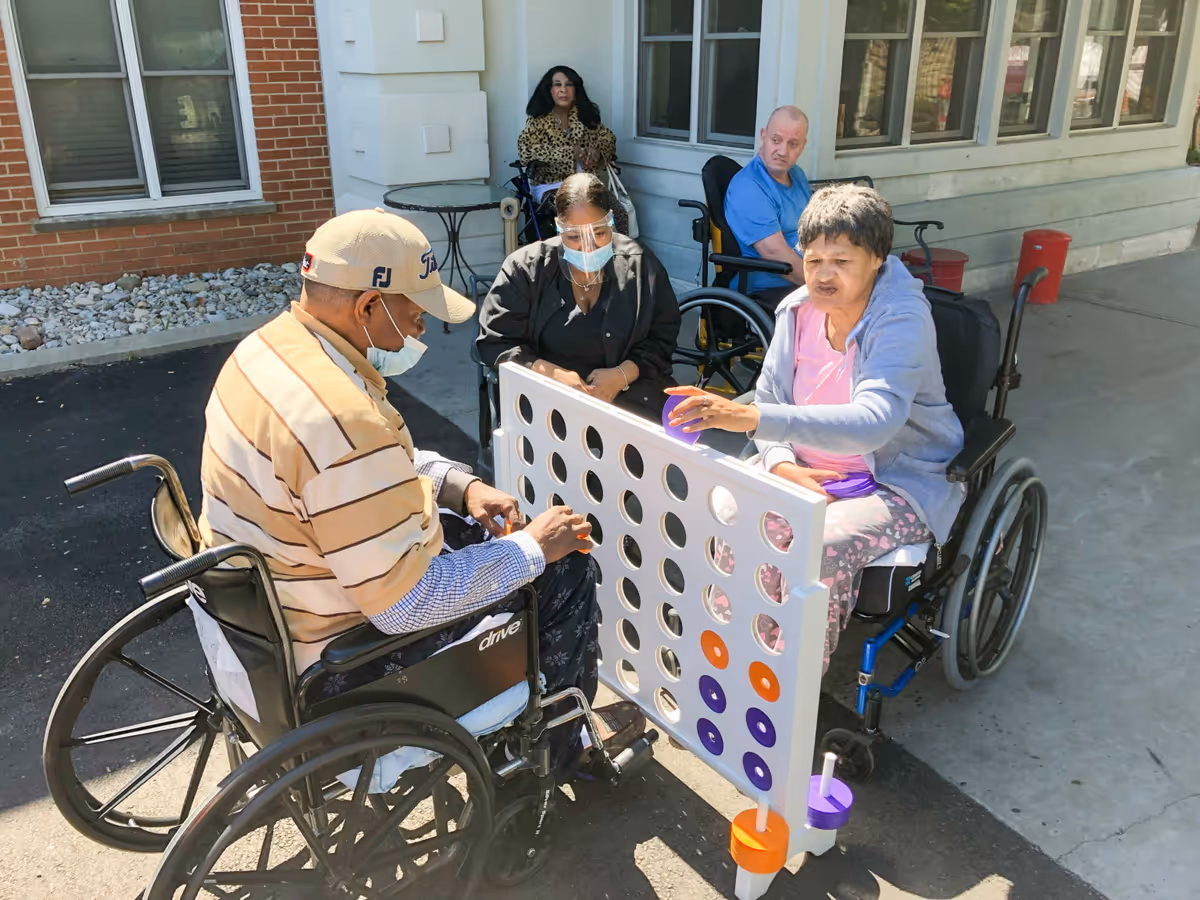Four elderly individuals, three in wheelchairs and one seated, are playing a large outdoor Connect Four game on a paved area outside a building with windows and brick walls. Two of the players are placing colored discs into the game board while the others watch. Some individuals are wearing face masks.
