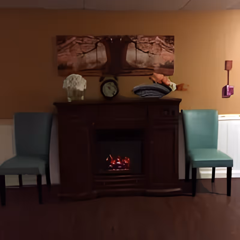 A cozy interior scene featuring a dark wooden fireplace with a small fire burning inside. On top of the fireplace mantel are decorative items including a clock and some sculptures. Two teal upholstered chairs are positioned on either side of the fireplace against a beige wall with white wainscoting. Above the mantel hangs a large abstract painting with earthy tones.