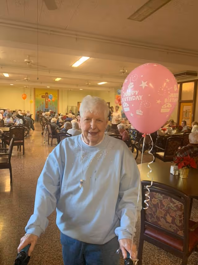 Elderly woman smiling in a busy dining room holding a pink "Happy Birthday" balloon.