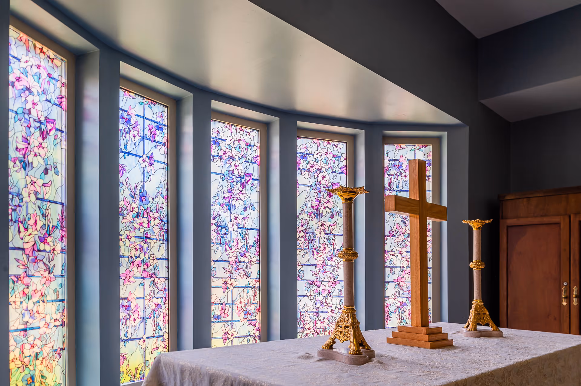 An altar with a wooden cross and two ornate golden candle holders in front of a row of tall stained glass windows featuring a floral design in shades of pink, purple, and blue.
