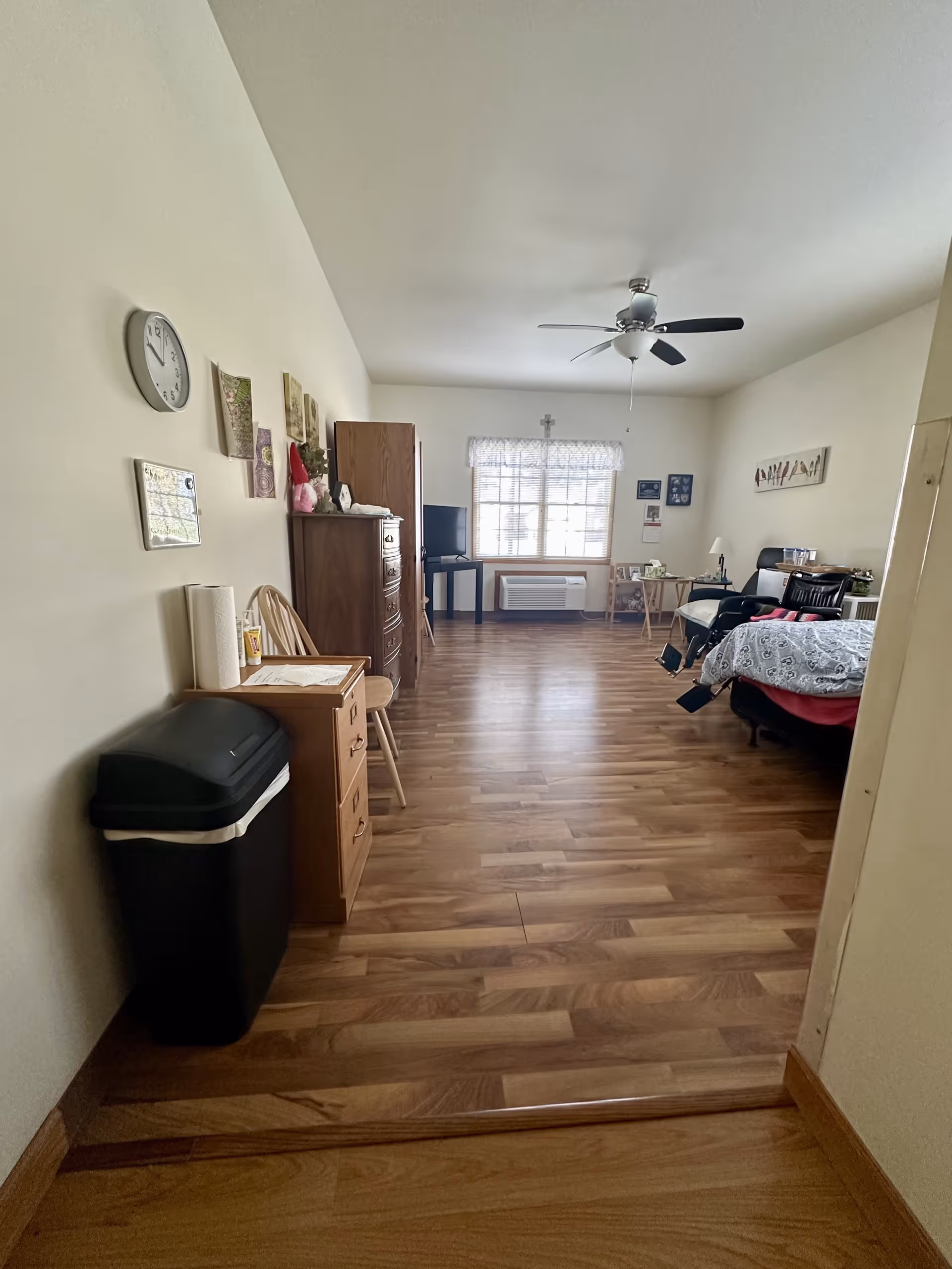A spacious senior living bedroom with wooden flooring, a ceiling fan, a bed with a patterned comforter, a black recliner chair, a wooden dresser, a small TV on a stand near a window with a lace curtain, and various wall decorations including a clock and framed pictures.
