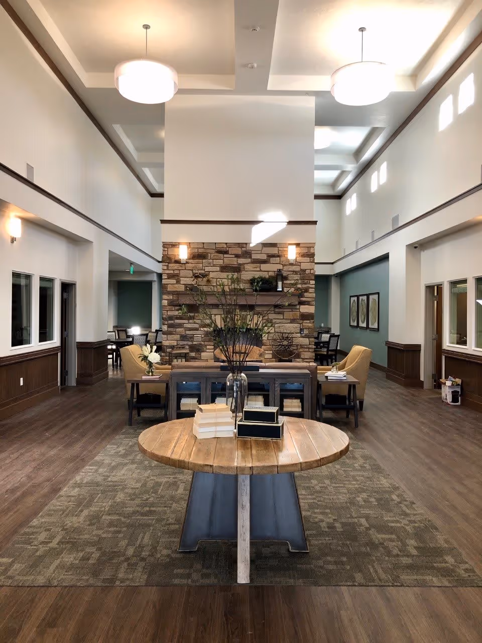 Bright, spacious assisted-living lobby with a round wooden table in the foreground, seating areas and a central stone fireplace under high ceilings.