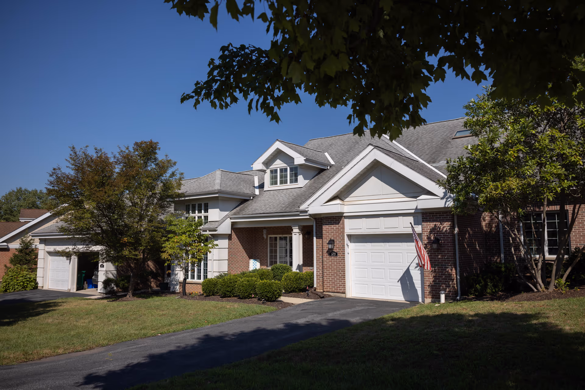 Exterior view of a residential-style building with a brick facade and white garage doors, surrounded by green trees and shrubs under a clear blue sky.