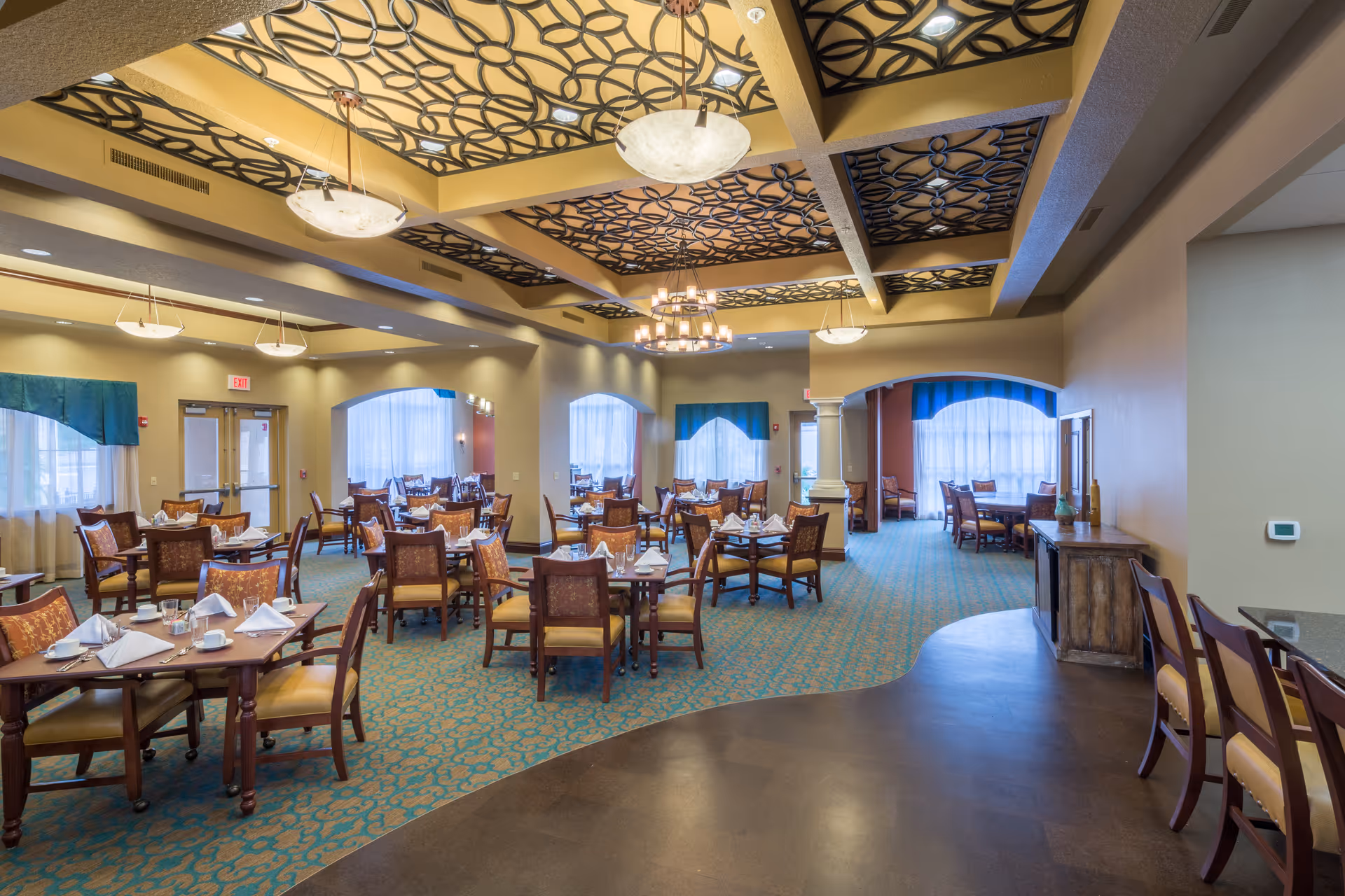 Spacious dining room with multiple tables and chairs arranged neatly, each table set with white napkins, cups, and glasses. The room features a decorative ceiling with patterned panels and hanging light fixtures. Large windows with sheer curtains allow natural light to fill the space. There is a curved counter area with chairs on the right side.