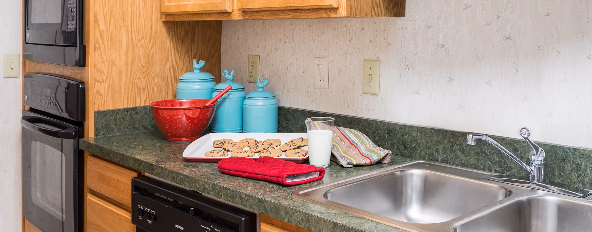 Kitchen countertop with a tray of chocolate chip cookies, a glass of milk, a red oven mitt, a striped cloth, and three turquoise canisters with rooster-shaped lids. The countertop is green with a double stainless steel sink and wooden cabinets. A black microwave and oven are built into the cabinetry on the left side.