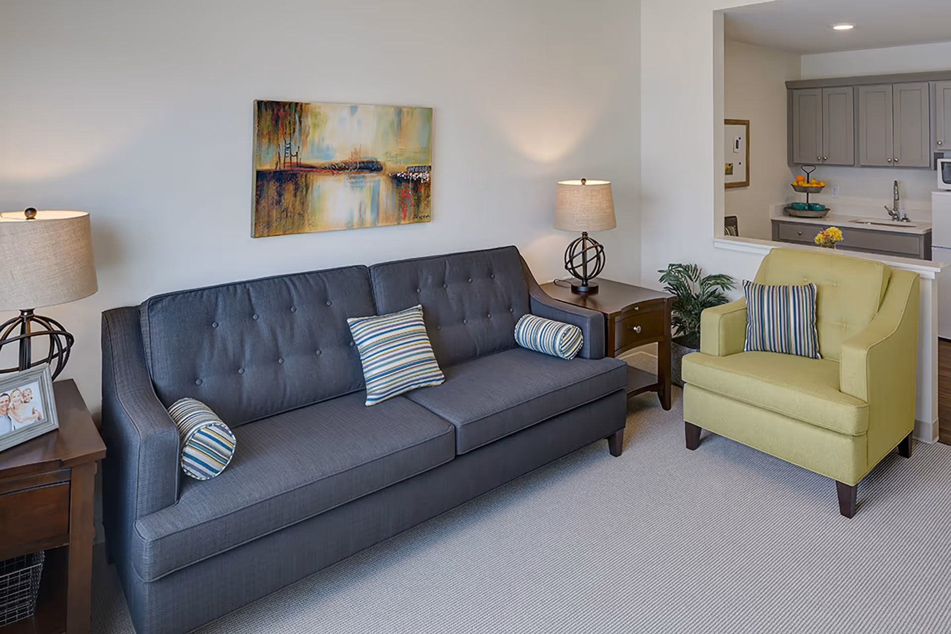A cozy living room with a dark gray sofa adorned with striped pillows and a light green armchair with a matching striped pillow. Two wooden side tables with lamps flank the sofa. A colorful abstract painting hangs on the wall above the sofa. In the background, a kitchen area with gray cabinets and a white countertop is visible through a pass-through window.