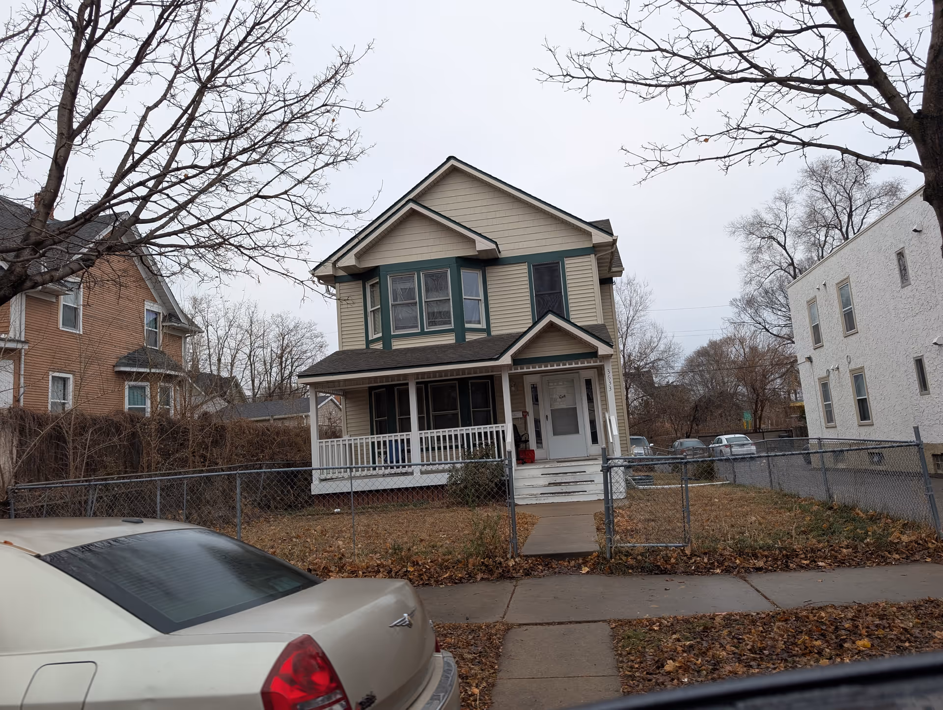 A two-story beige house with green trim and a covered front porch, surrounded by a chain-link fence. There are leafless trees and a beige car parked in front on a cloudy day.