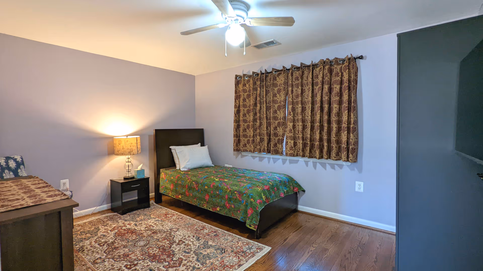 A simple bedroom with a single bed covered with a green floral bedspread, a white pillow, a nightstand with a lamp and tissue box, a patterned area rug on wooden floor, a window with brown patterned curtains, and a ceiling fan with light.