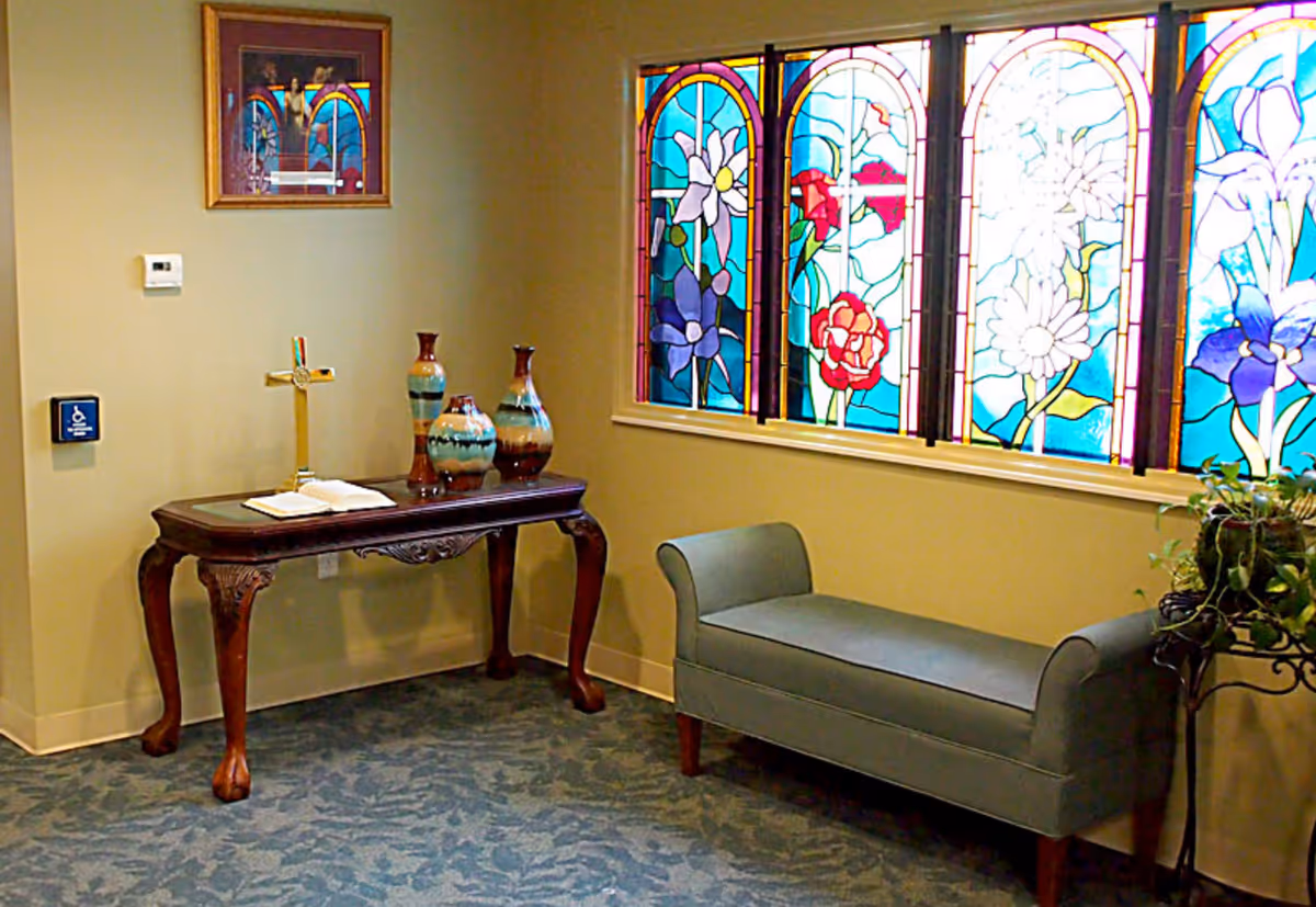 A quiet room with a decorative wooden table holding a cross, an open book, and three vases. To the right, there is a cushioned bench and a plant stand with a green plant. The wall features colorful stained glass windows with floral designs.