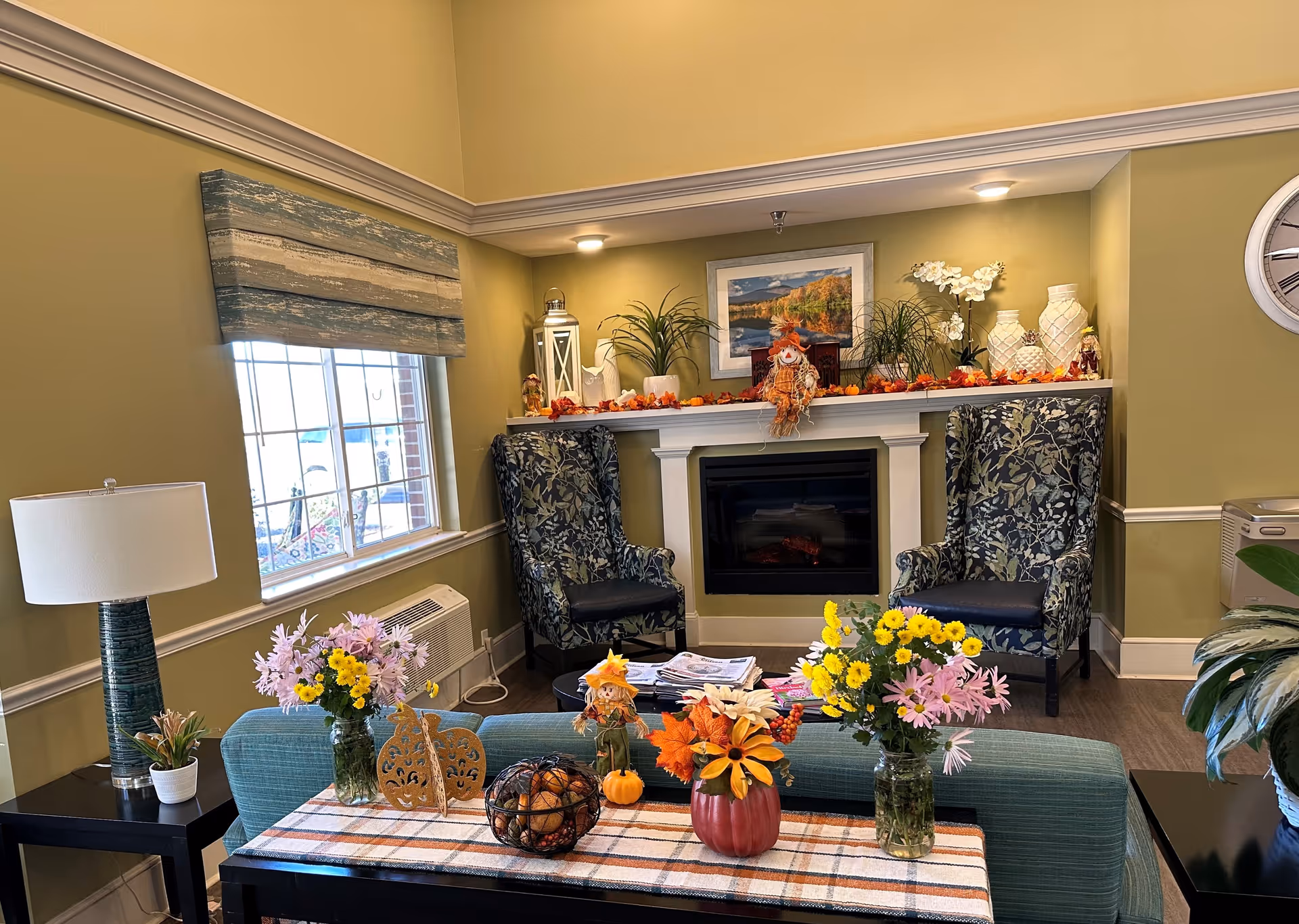 Cozy senior living room with a fireplace flanked by two patterned armchairs, a table with flowers and seasonal decor, and a window letting in daylight.