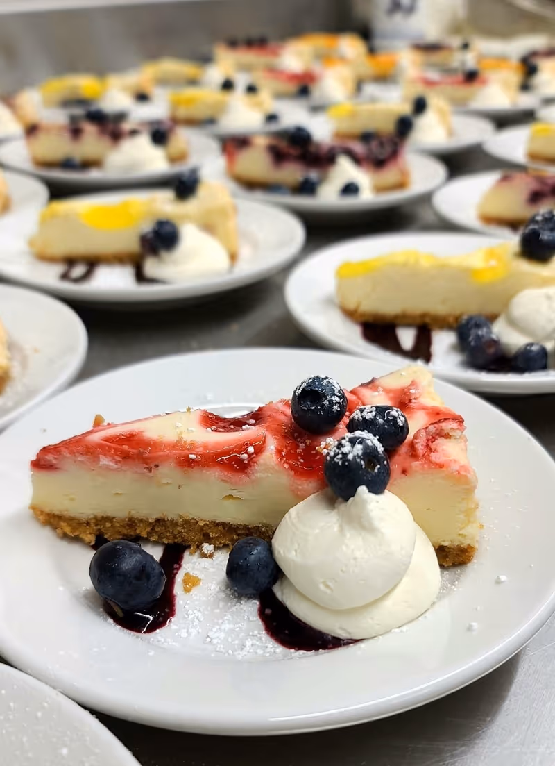 Close-up view of multiple plates with slices of cheesecake topped with blueberries, whipped cream, and a drizzle of berry sauce, arranged on a table.