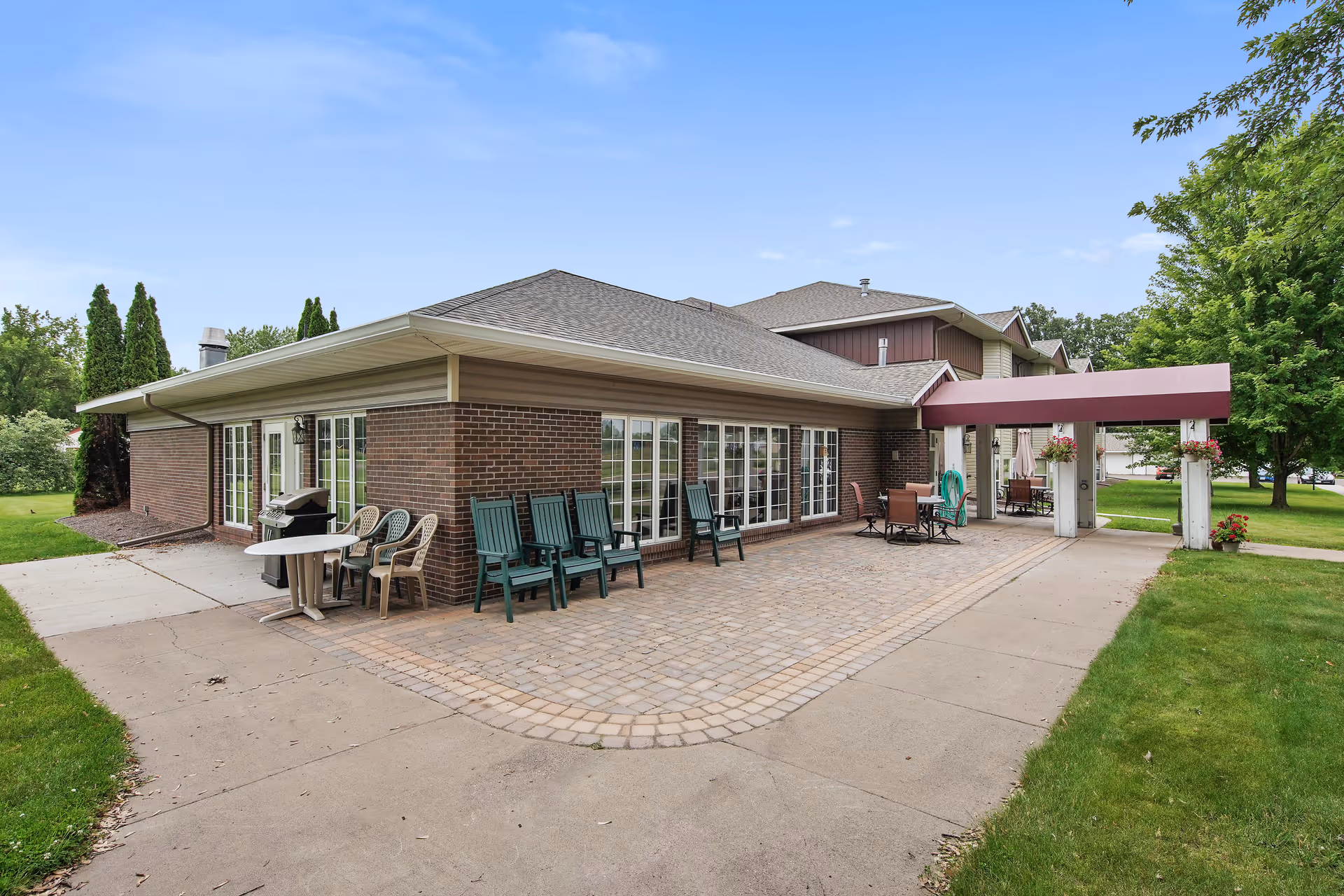 Exterior view of a single-story brick senior living building with a paved patio, outdoor chairs, and a covered entrance canopy.
