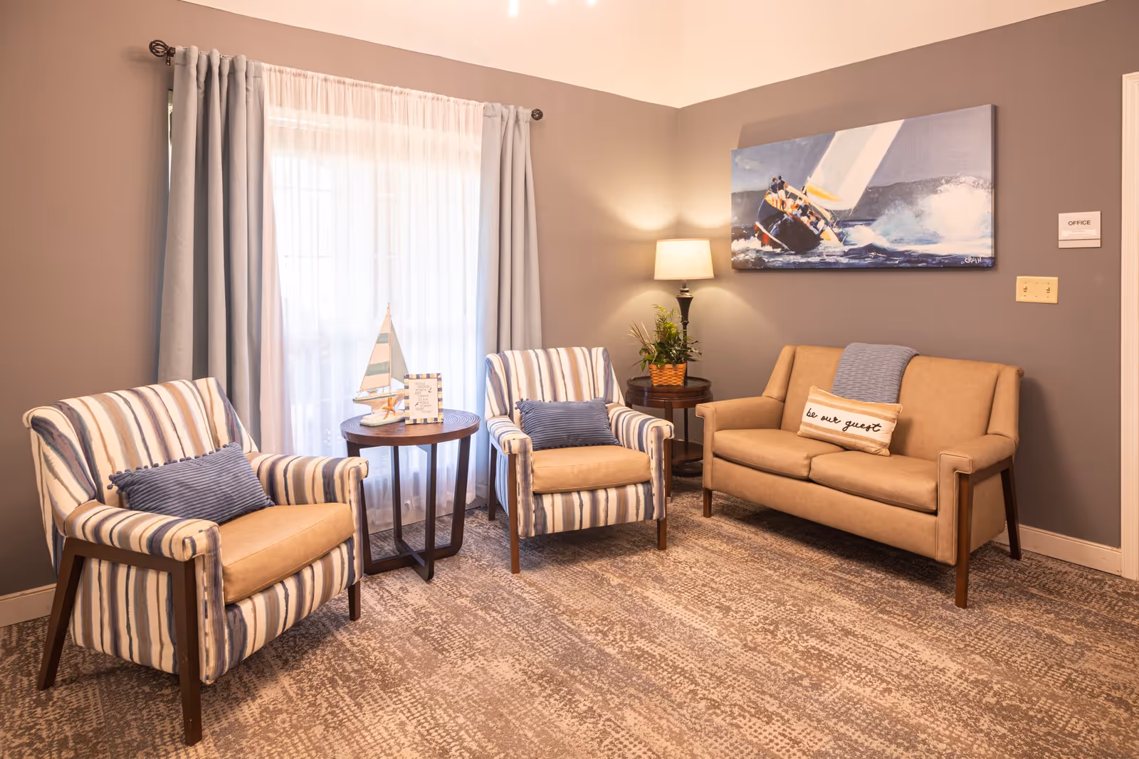 A cozy sitting area with two striped armchairs, a beige loveseat, side tables, a lamp, and nautical artwork by a curtained window.