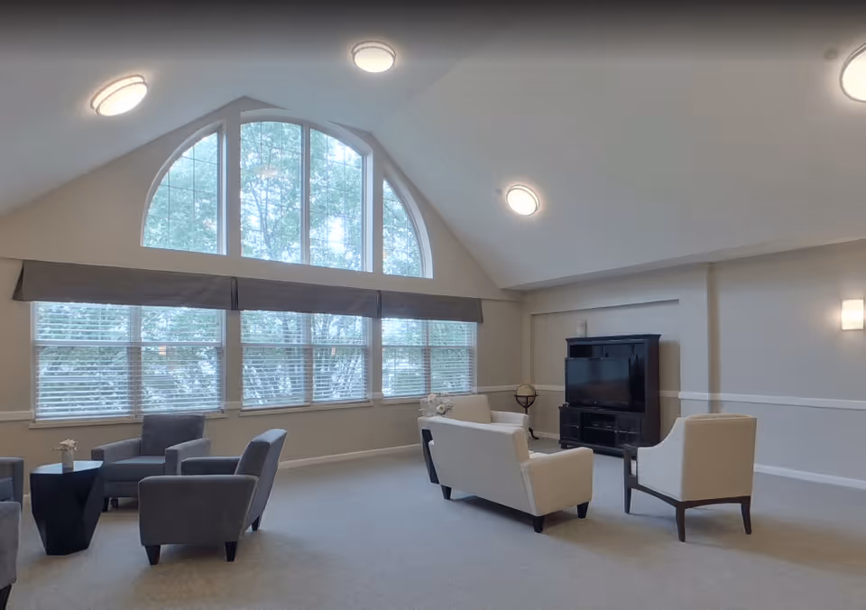 A spacious living room with large arched windows letting in natural light. The room features two white armchairs and a white loveseat arranged around a black TV stand with a flat-screen television. There are also three gray armchairs around a small black table. The walls are painted light gray with white trim, and the ceiling has recessed lighting fixtures.