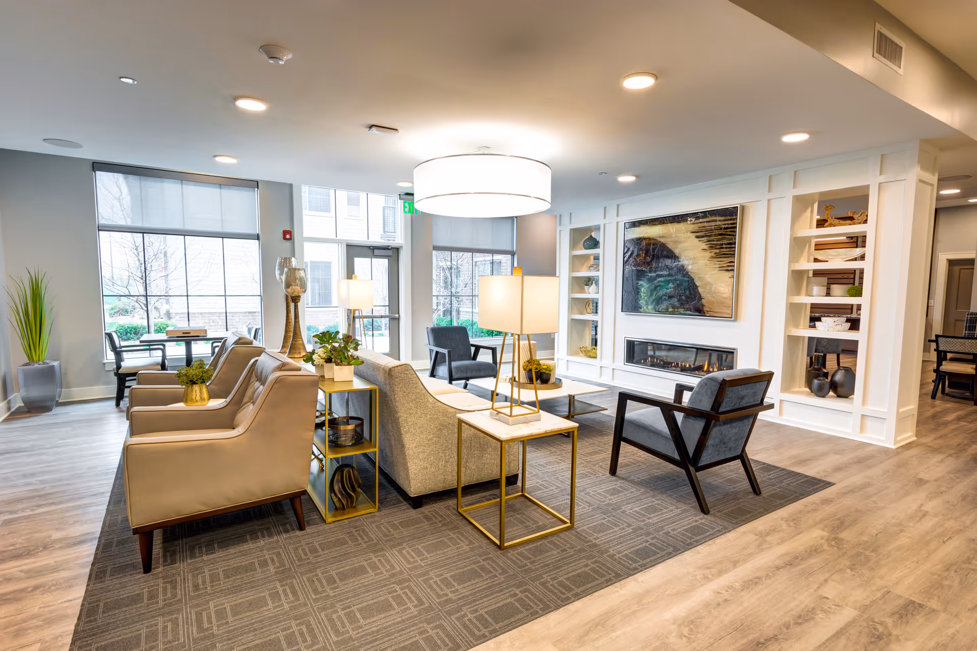 Bright, modern senior living common area with sofas, armchairs, side tables, built-in shelving and a fireplace beneath wall art.