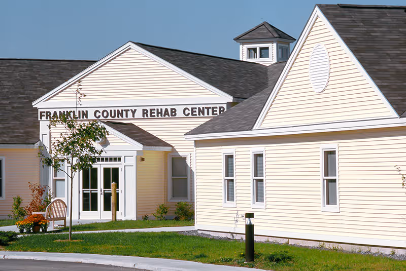 Exterior view of the Franklin County Rehab Center building with light yellow siding, multiple windows, and a main entrance with double glass doors. There is a small tree and a bench near the entrance, with a clear blue sky in the background.