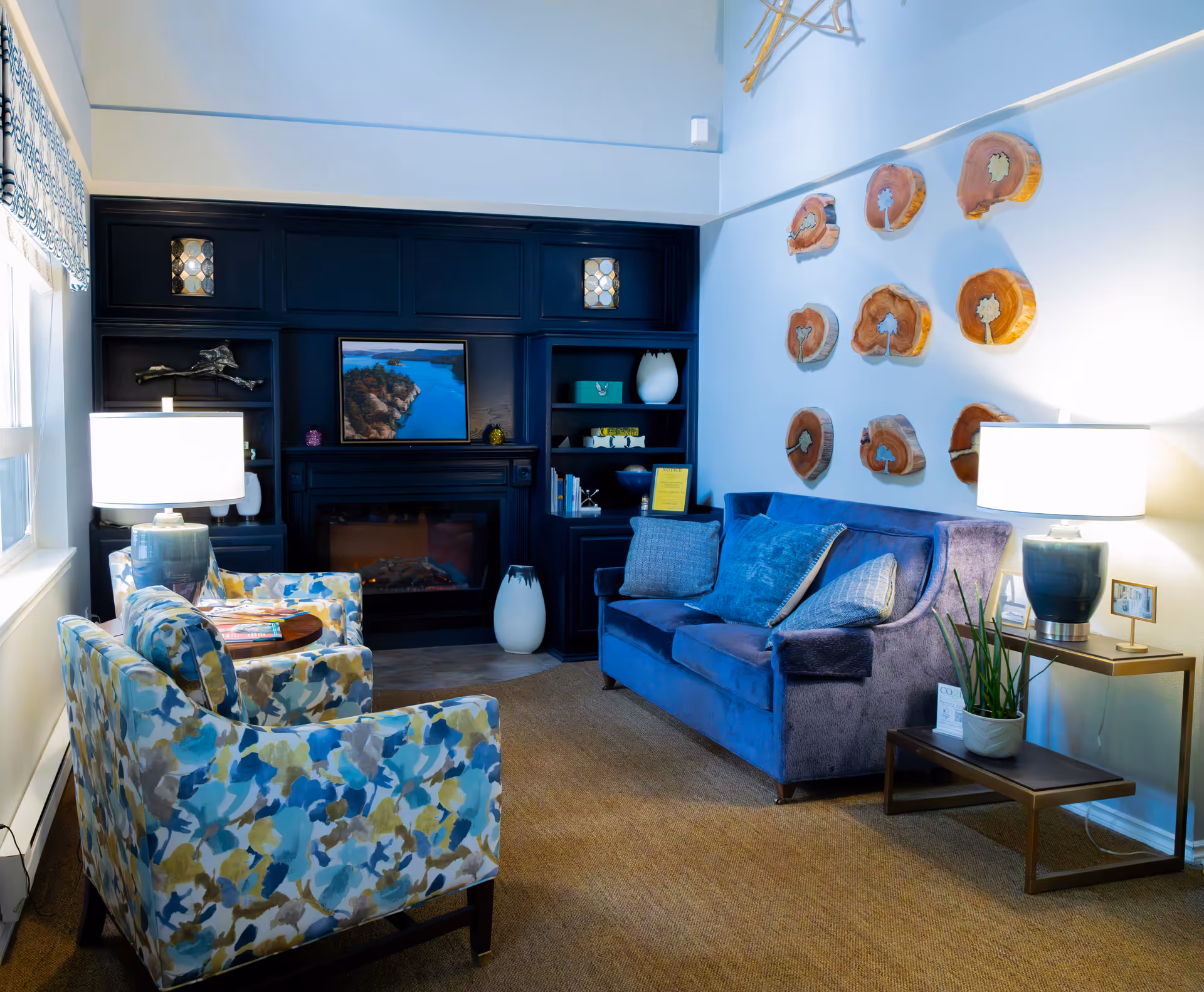 A cozy living room with a blue velvet sofa and two floral-patterned armchairs around a small round wooden table. The room features a dark built-in shelving unit with a fireplace and decorative items, including a framed picture of a coastal scene. Two table lamps with white shades provide warm lighting. The wall above the sofa is decorated with wooden art pieces resembling tree cross-sections. A small side table with a potted plant is next to the sofa.