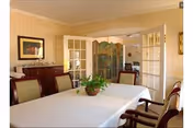 A formal dining room with a long table covered by a white tablecloth, chairs around it, a potted centerpiece, sideboard, and French doors leading to another room.