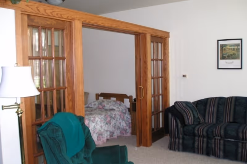 Living room with green upholstered seating and a floor lamp opening through wooden glass-paneled sliding doors to a bedroom with a floral bedspread.