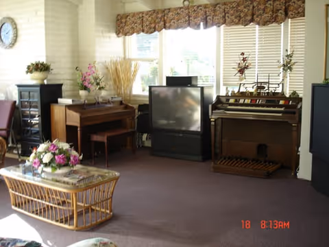 A cozy living room with a large window covered by floral valances, featuring a vintage television set, a piano, an organ, a glass-top coffee table with a floral arrangement, and various decorative plants and flowers.
