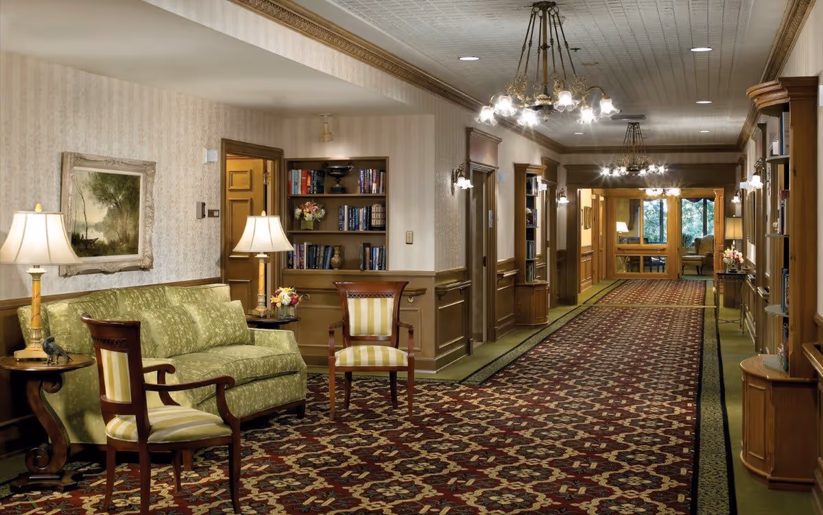 A long, carpeted hallway in a senior living facility with patterned red and beige carpet, beige wallpaper, and wood paneling. The hallway is furnished with a green patterned sofa, two wooden chairs with striped cushions, side tables with lamps, built-in bookshelves filled with books and decorative items, and multiple chandeliers and wall sconces providing warm lighting. At the end of the hallway, large windows reveal greenery outside.