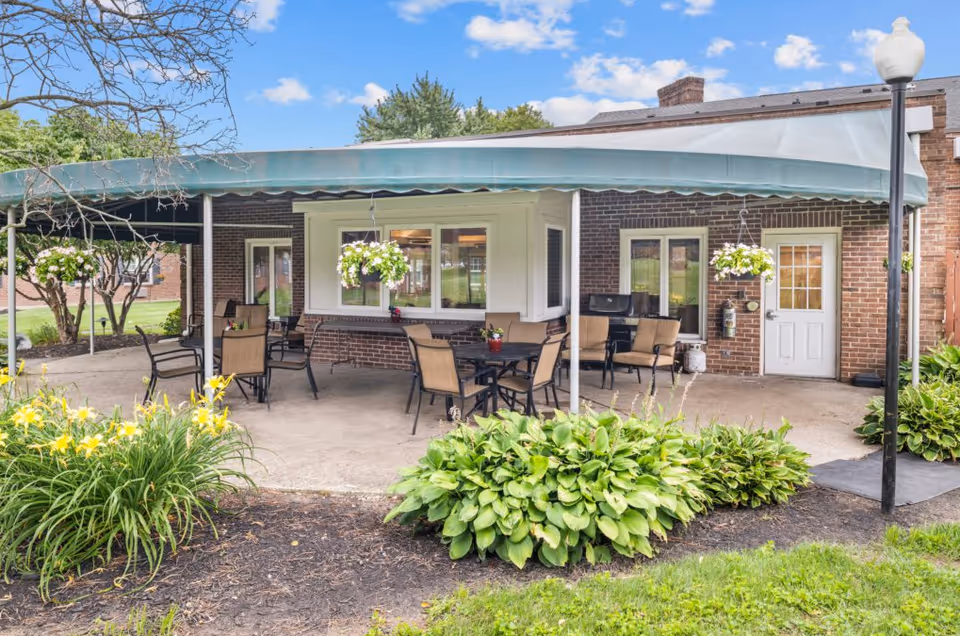Outdoor covered patio area at Oakleaf Village of Columbus with several tables and chairs, hanging flower baskets, and surrounding greenery including yellow flowers and leafy plants. The patio is attached to a brick building with windows and a white door.