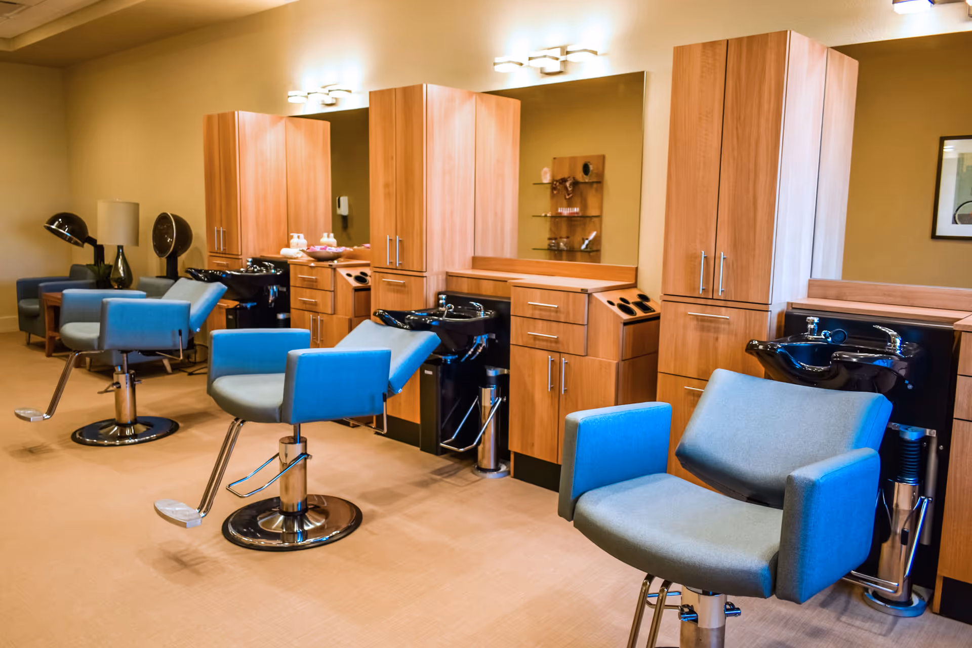 Interior view of a salon area with multiple blue salon chairs in front of wooden cabinets and black hair washing sinks. The room has warm lighting and a beige carpeted floor.