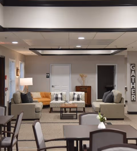 A cozy living room area in an assisted living facility with beige and orange sofas, patterned armchairs, a wooden coffee table, and a lamp. The walls are light-colored with a 'GATHER' sign on the right side. There are tables and chairs in the foreground, and a door and a small cabinet with decorative items in the background.