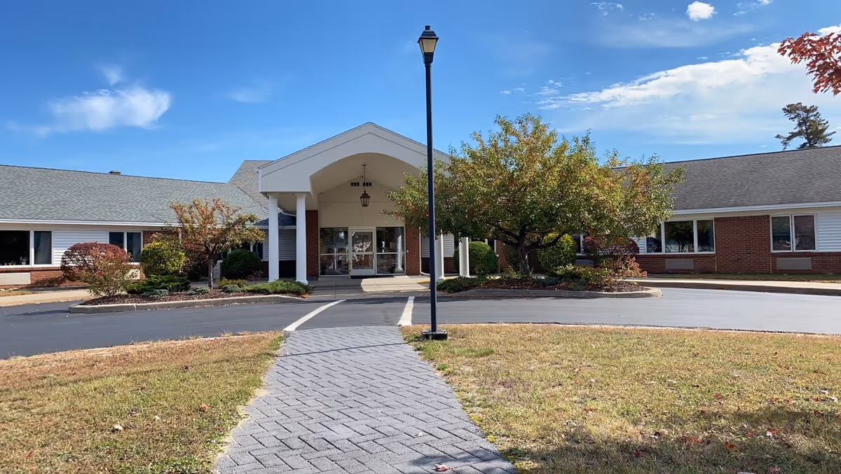 Front exterior view of a single-story senior living community building with a covered entrance supported by white columns, a paved walkway leading to the entrance, a streetlamp in the center, and trees and shrubs around the building under a partly cloudy blue sky.