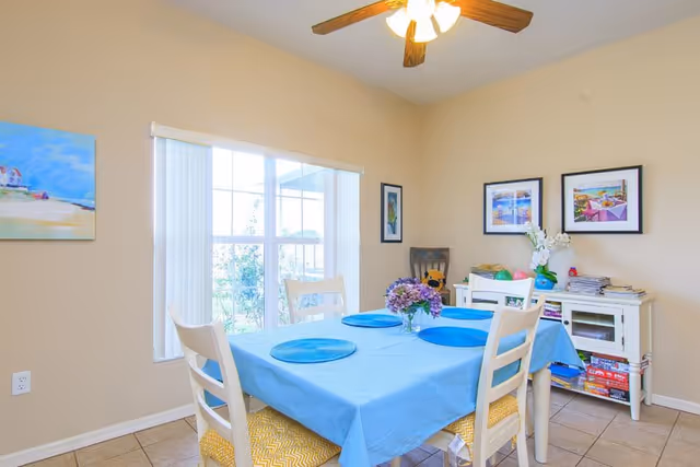 Bright dining room with a blue tablecloth and four chairs under a ceiling fan beside a large window and a sideboard with decor.