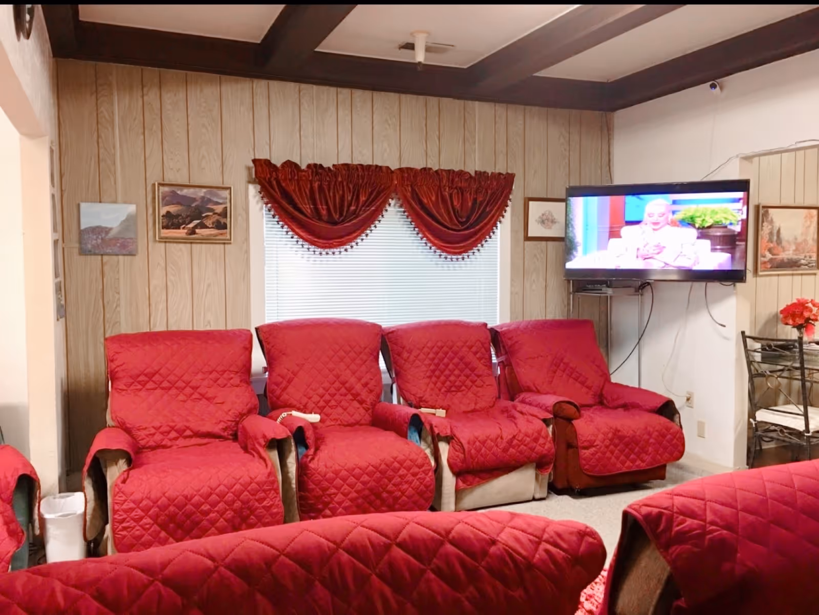 A cozy living room with four recliner chairs covered in red quilted fabric facing a wall-mounted flat-screen TV. The walls are wood-paneled with several framed paintings and a window with red valance curtains. A small dining table with chairs is partially visible on the right side.