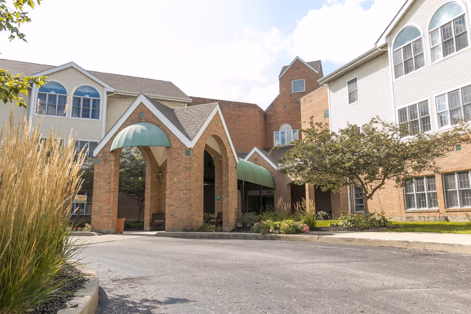 Exterior view of Riverview - An Optalis Health & Rehabilitation Center showing a brick building with white siding, multiple windows, and a covered entrance with green awnings. There are plants and trees around the entrance and a paved driveway in front.