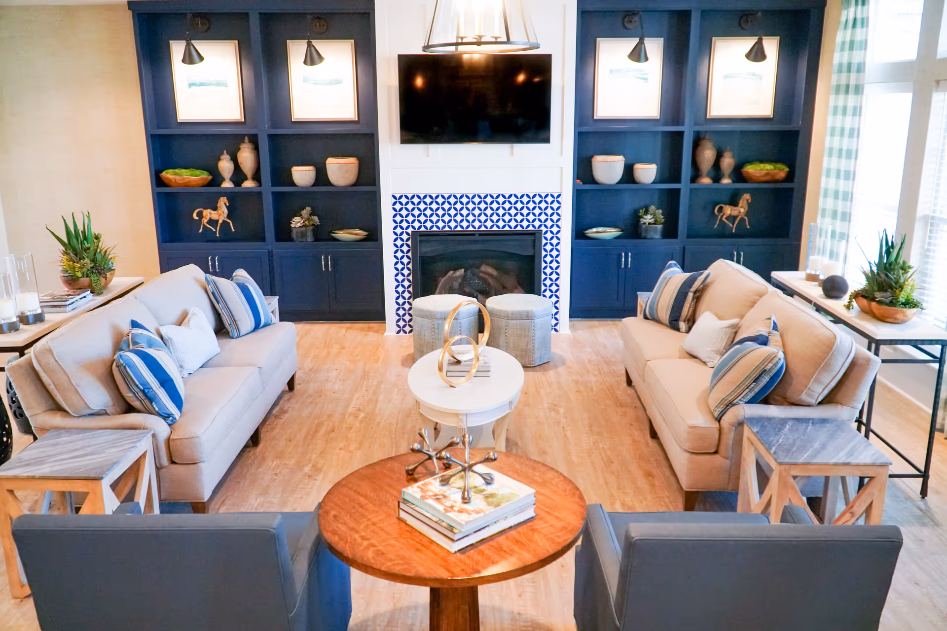 A bright and cozy living room with two beige sofas adorned with blue and white striped pillows facing each other. Between them is a small round white table with decorative items. In front of the sofas are two gray armchairs and a round wooden coffee table with books and a decorative piece. The room features a fireplace with blue and white patterned tiles, flanked by built-in navy blue shelves holding vases, bowls, and horse figurines. A flat-screen TV is mounted above the fireplace. Large windows with blue and white checkered curtains allow natural light to fill the space.