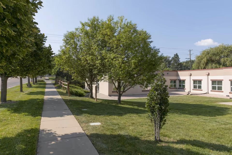 Sidewalk lined with trees and grass leading alongside a single-story building with multiple windows under a clear blue sky.