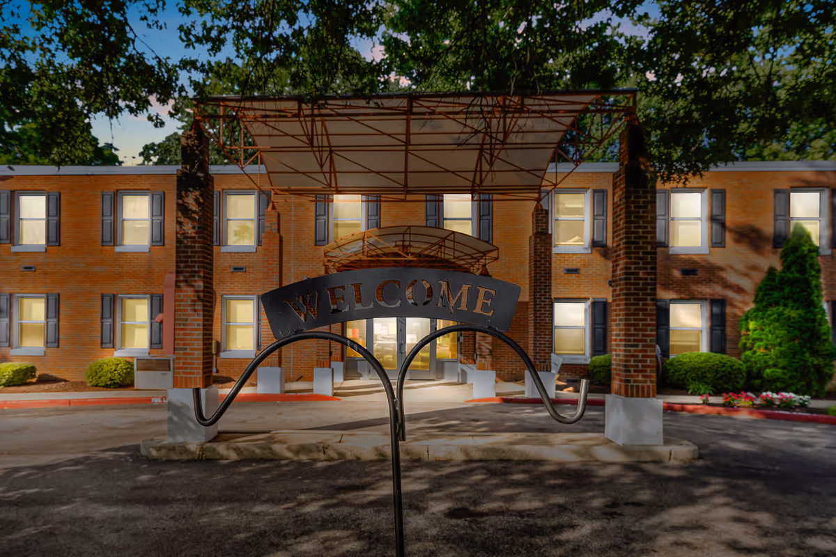Exterior front view of a two-story brick building with multiple windows, a covered entrance supported by brick pillars, and a metal sign in the foreground that reads 'WELCOME'. The scene is set during dusk with lights on inside the building and trees framing the top of the image.