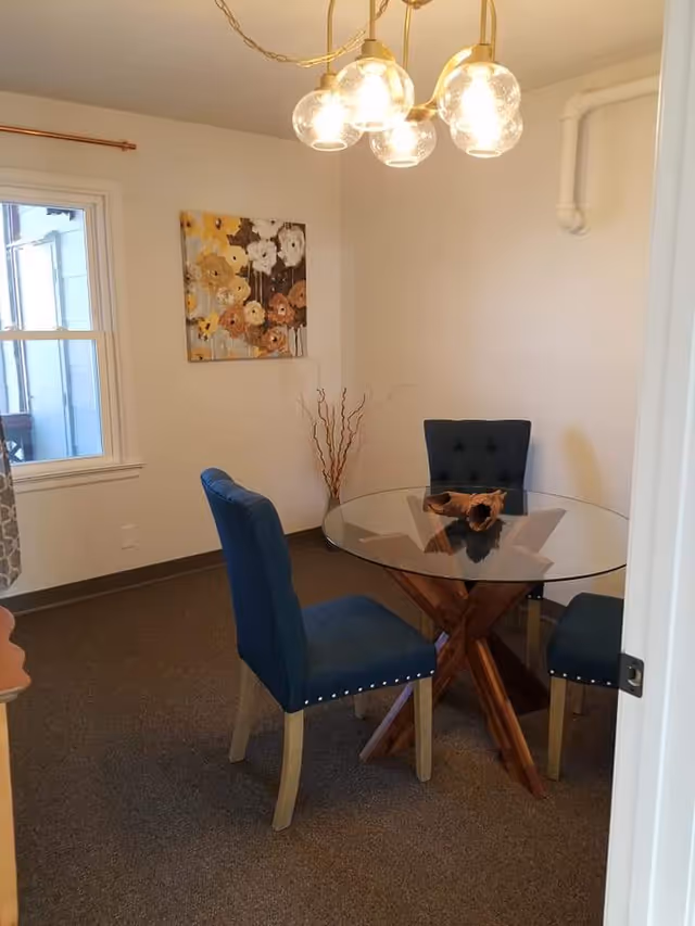 A small dining area with a round glass table supported by a wooden base and three blue upholstered chairs. A modern chandelier with five glass globe lights hangs above the table. There is a window with a curtain on the left wall and a floral painting on the wall behind the table. A decorative branch arrangement is placed in the corner.