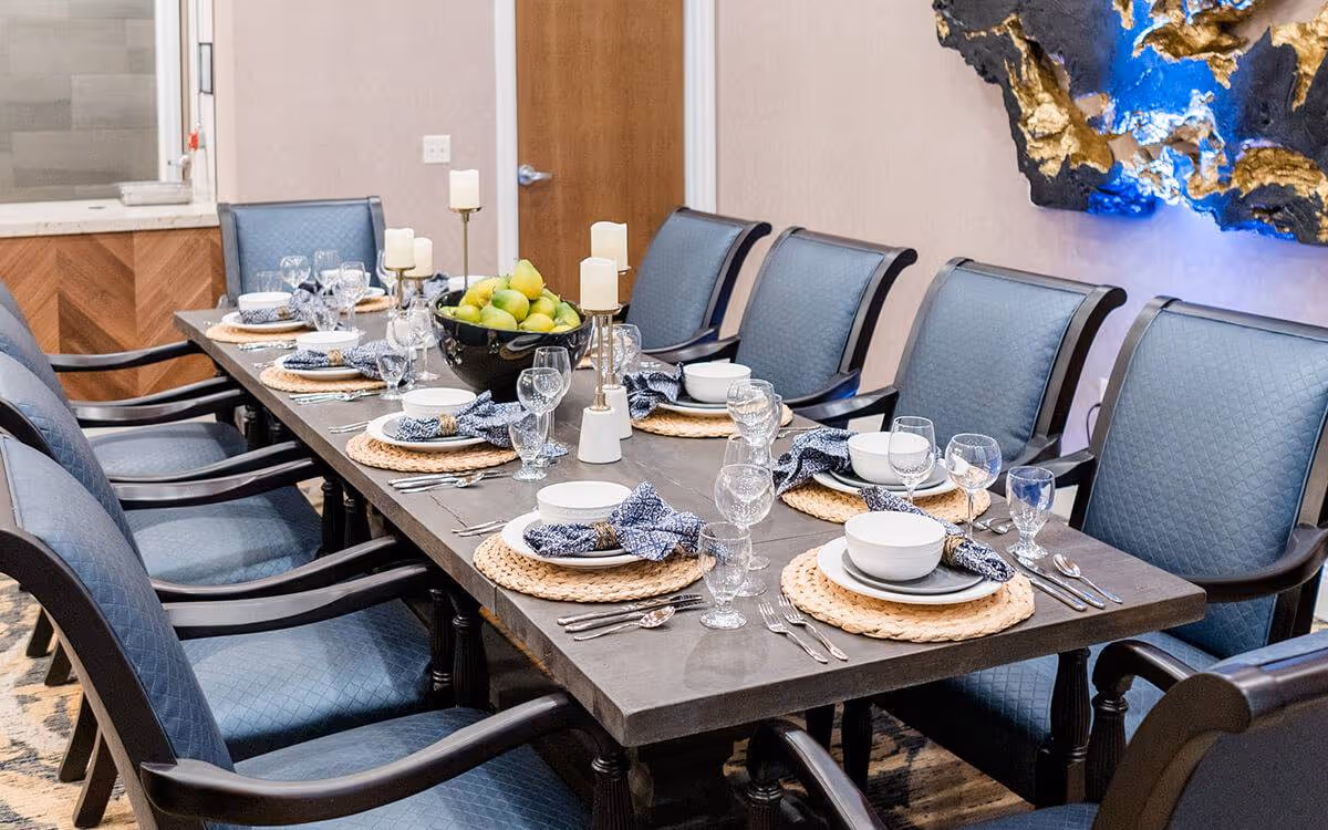 A dining table set for eight with blue cushioned chairs around it. The table is decorated with woven placemats, white plates, bowls, silverware, glassware, blue patterned napkins, three candle holders with white candles, and a black bowl filled with green apples. The room has beige walls, a wooden door, and a decorative wall art piece with blue and gold accents.