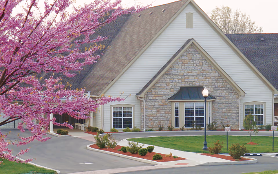 Front exterior of a senior living building with a stone-faced entrance, blooming pink tree, and a circular driveway.