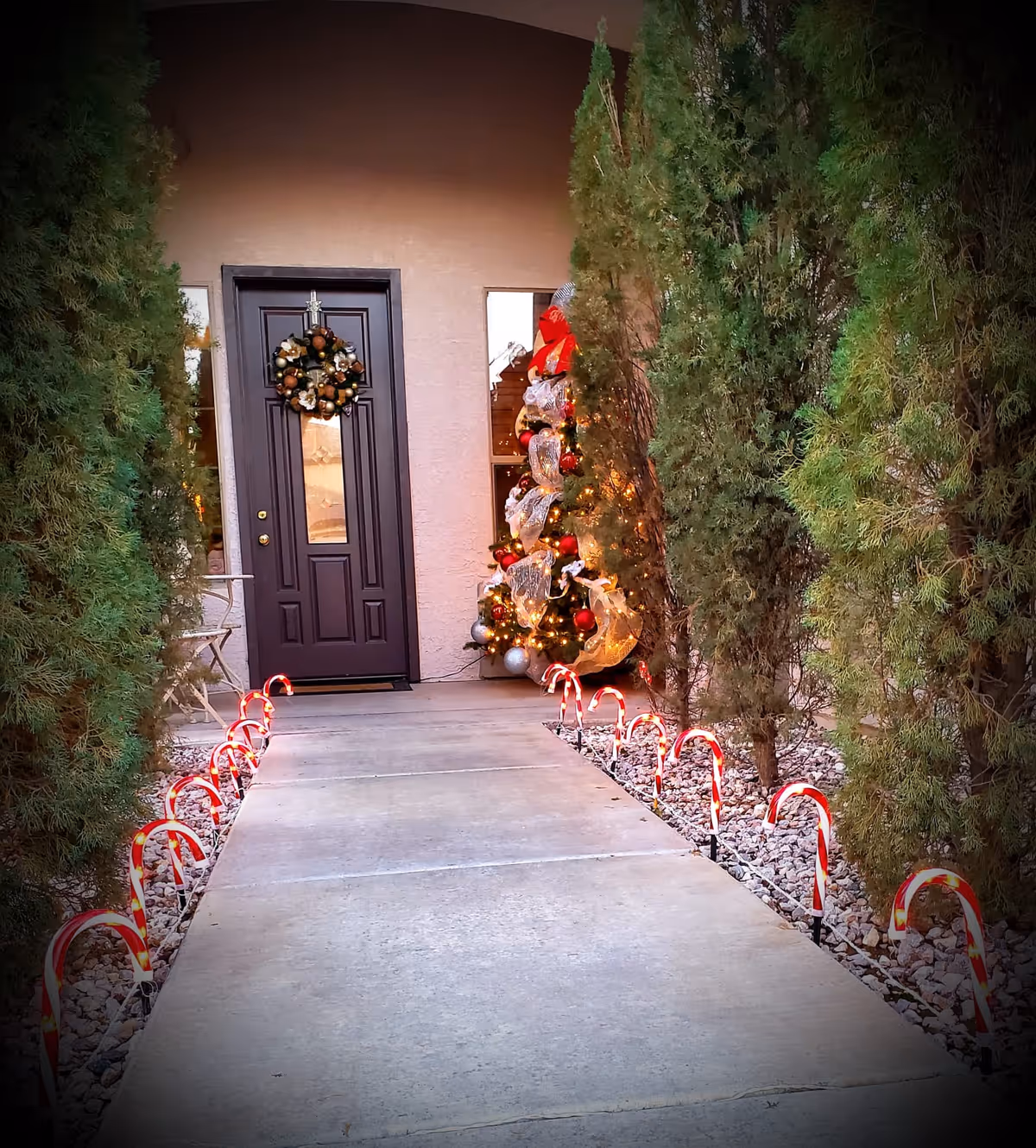 A walkway lined with small candy cane lights leads to a dark front door decorated with a Christmas wreath. To the right of the door, there is a Christmas tree adorned with lights, ribbons, and ornaments. Tall green bushes flank both sides of the walkway.