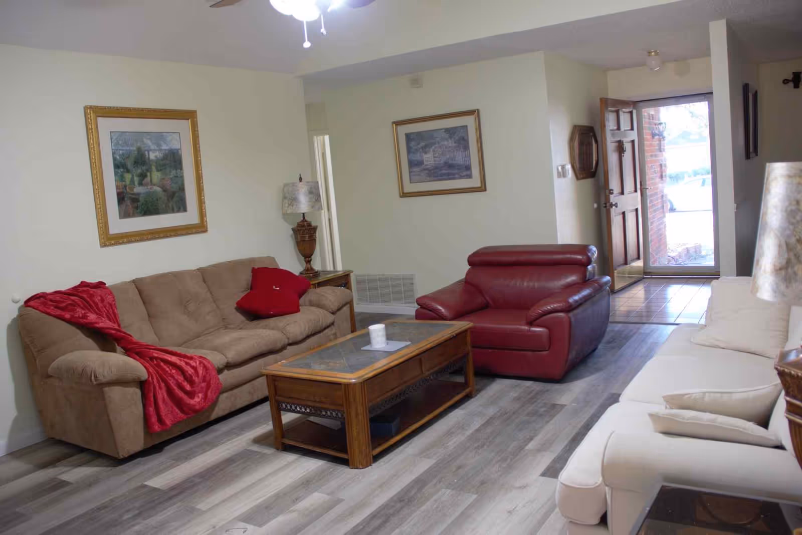 A cozy living room with a beige couch draped with a red blanket and red pillows, a red leather armchair, a white couch with beige pillows, a wooden coffee table with a candle on it, two framed paintings on the walls, a wooden side table with a decorative lamp, and an open front door showing a glimpse of the outside.