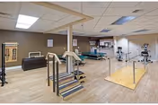 A spacious physical therapy or rehabilitation room with various exercise and therapy equipment including parallel bars, stairs, exercise bikes, and therapy tables. The room has wood flooring, beige walls, and a drop ceiling with fluorescent lighting.