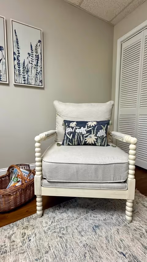 A cozy beige armchair with a decorative floral pillow is placed on a patterned rug in a room. To the left of the chair is a wicker basket containing magazines. On the wall behind the chair are two framed botanical prints featuring lavender flowers. A white louvered closet door is visible on the right side of the image.