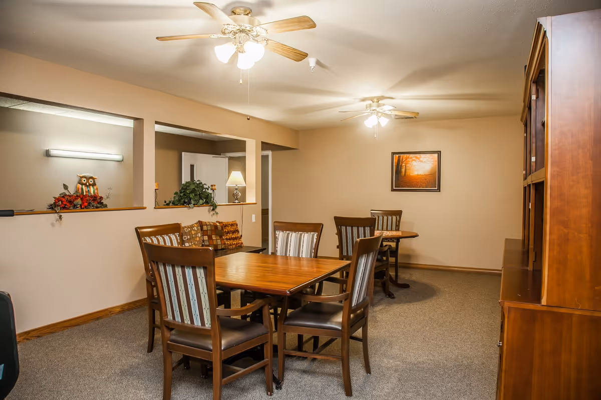 A cozy interior room with two wooden tables surrounded by chairs with striped upholstery. The room has beige walls, carpeted floor, two ceiling fans with lights, a wooden cabinet on the right, and a framed picture of an autumn forest on the far wall. There are decorative plants and an owl figurine on a ledge near a doorway with a lamp in the background.