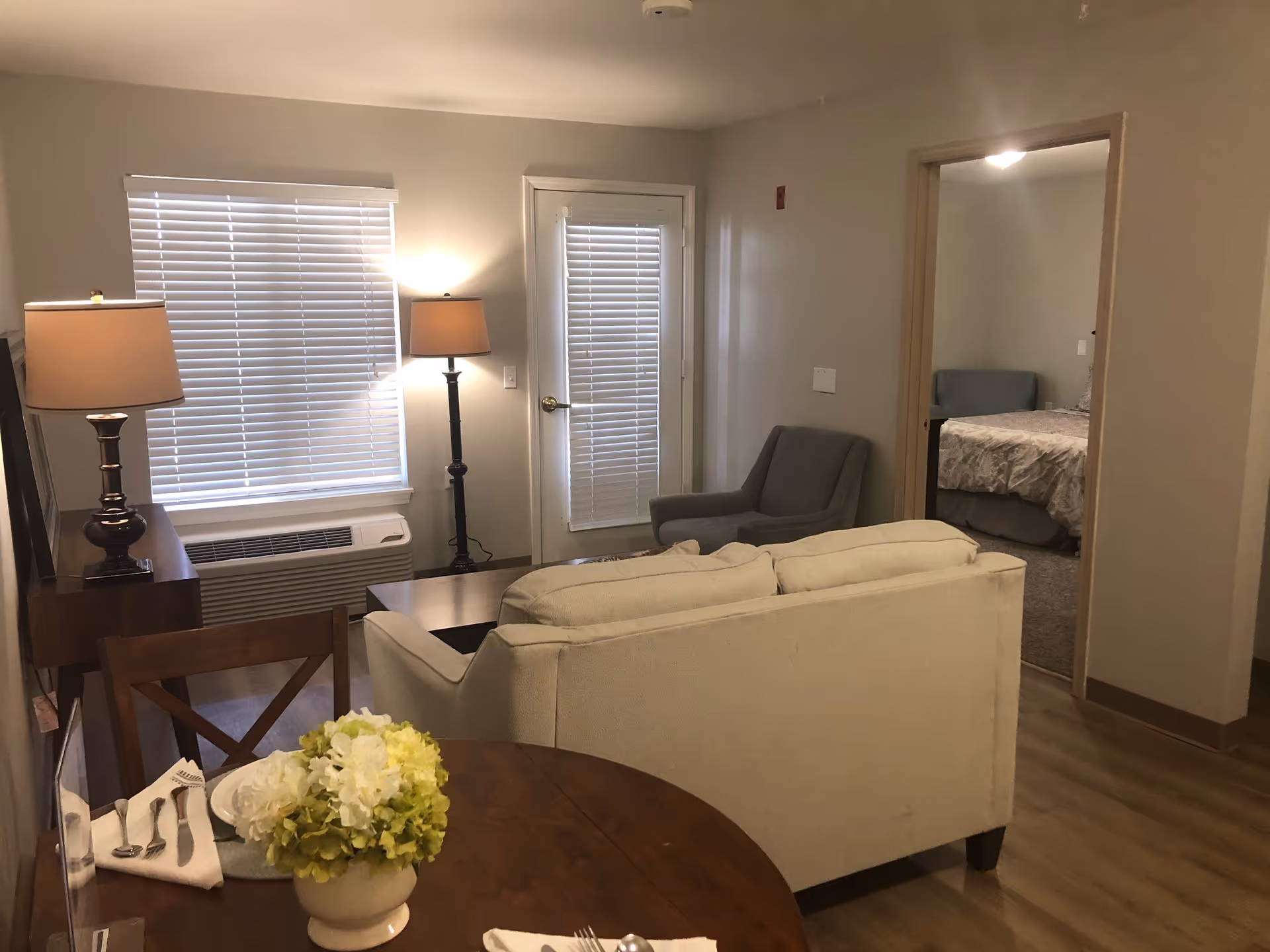 Interior view of a senior living apartment showing a small living room with a beige couch, a gray armchair, two lamps, a window with blinds, and a door with blinds. A round wooden dining table with a flower arrangement and place settings is in the foreground. A bedroom with a bed and a blue chair is visible through an open doorway.