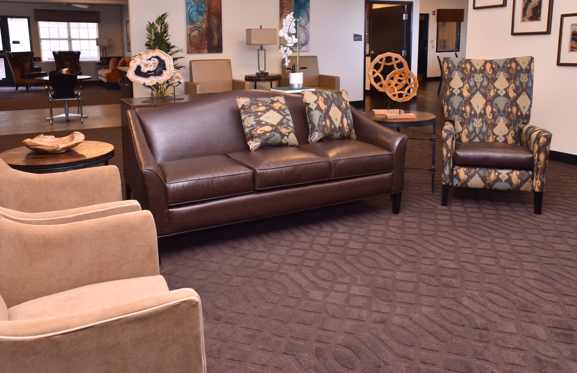 A cozy seating area in a senior living facility featuring a brown leather sofa with patterned cushions, two beige armchairs, and a patterned armchair. The room has a brown carpet with a geometric design, side tables with decorative items, and artwork on the walls. In the background, there are more seating areas and windows letting in natural light.